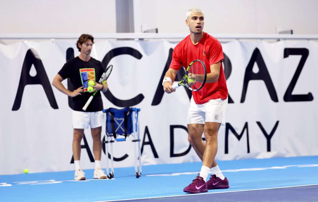 Juan Carlos Ferrero stands behind and watches Carlos Alcaraz prepare to serve